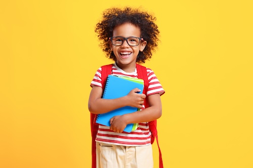 Adorable diligent little black schoolboy with Afro hair in casual clothes and eyeglasses laughs and looking at camera while standing against yellow background with notebooks