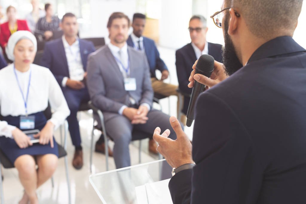 Rear view of male mixed race speaker speaks with microphone to diverse business people in a business seminar in a conference room