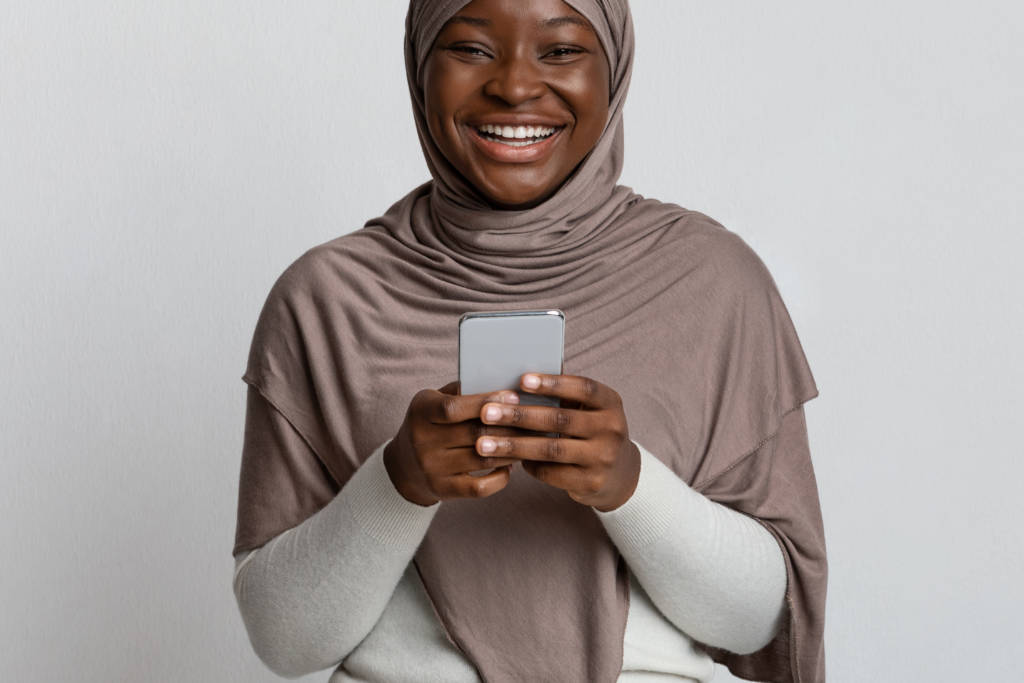 Joyful Black Muslim Lady In Hijab Holding Smartphone And Laughing At Camera