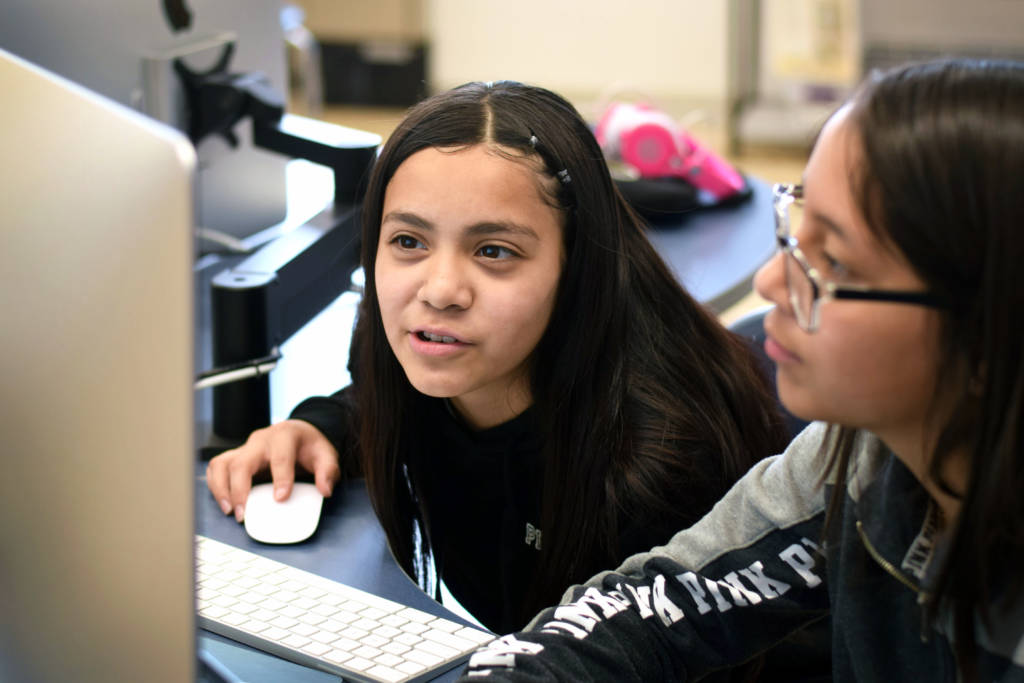 Two young students work together at a computer in a classroom setting.