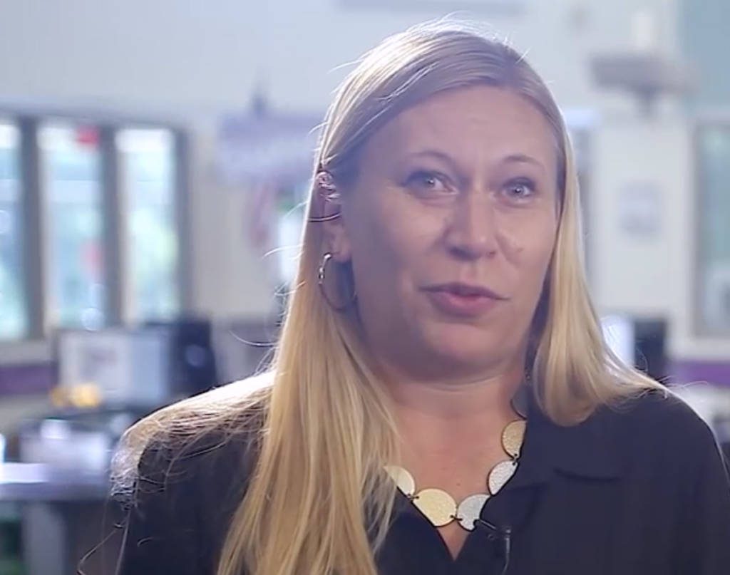 Woman with long blonde hair wearing a black blazer and necklace in an office setting.
