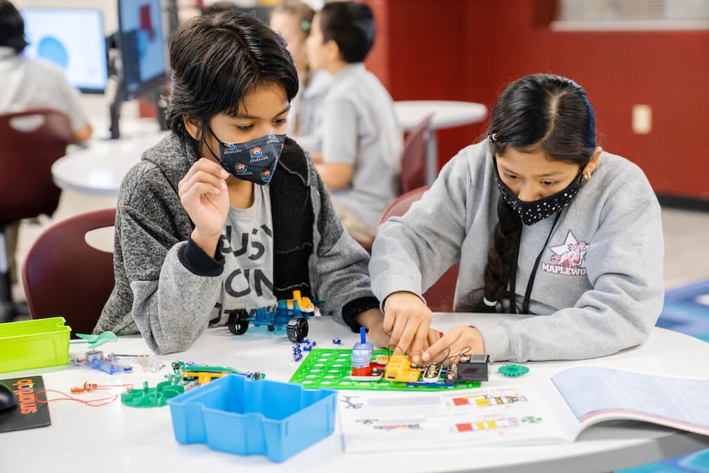 a young boy and girl are sitting at a table, with masks on. They are working together to build something using instructions.