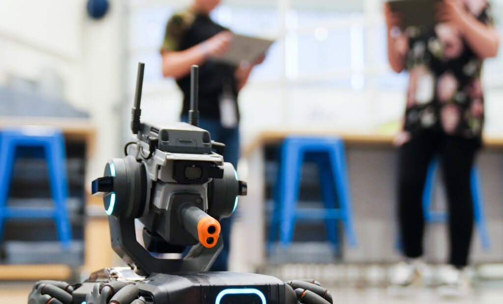 photo of a black metal robots head with two ear-like antennae and two people blurred in the background using ipads to control the bot