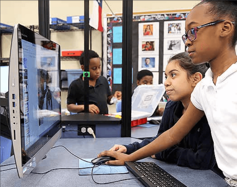 Two elementary students work together on a computer while classmates collaborate on a project-based learning assignment in the background.