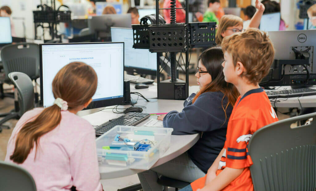three school aged kids share a computer in a computer lab, playing and learning STEM together