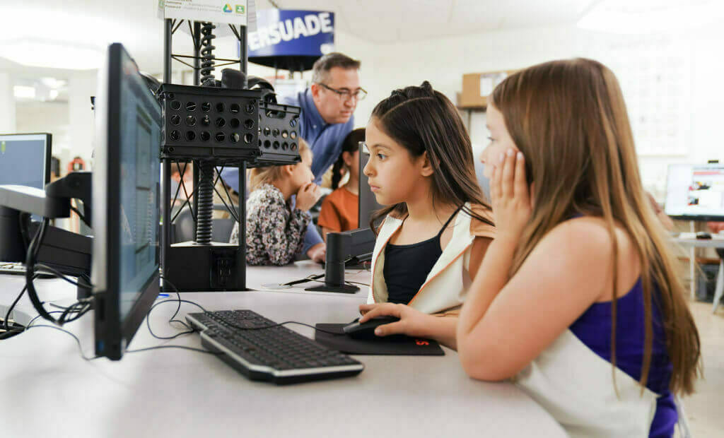 two girls with long straight hair look at a computer screen together, one girl is controlling the mouse attached to the computer with her hand