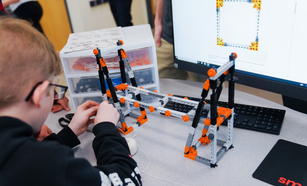 boy with light hair and glasses builds a bridge with lego-like toys in a STEM classroom setting