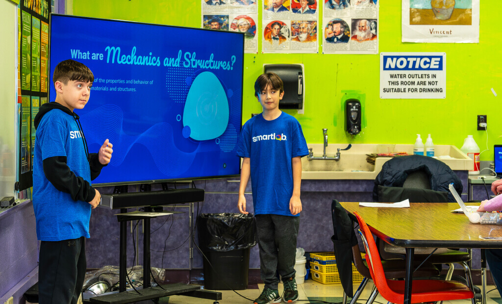 two middle school boys with dark hair and blue smartlab tshirts stand near a computer screen showing a slide about mechanics and structures in a smartlab classroom setting