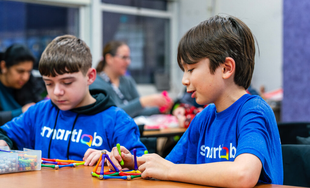 two boys in blue tshirts sit together at a table in a classroom and build structures using magnetic sticks