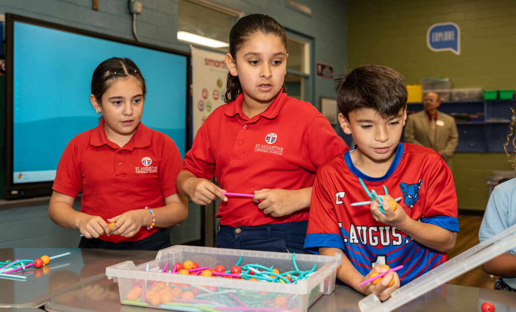 three school aged children in red shirts stand together at a table in a classroom setting while playing with engineering-inspired materials