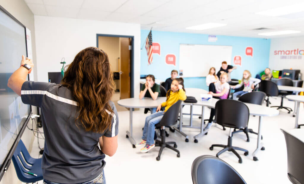 A woman stands at the front of a classroom, teaching a diverse group of students seated at desks.