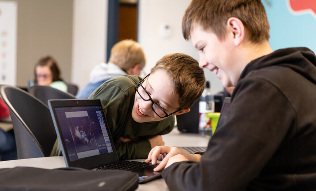 Two boys are focused on a laptop computer while seated at a desk in a classroom setting.