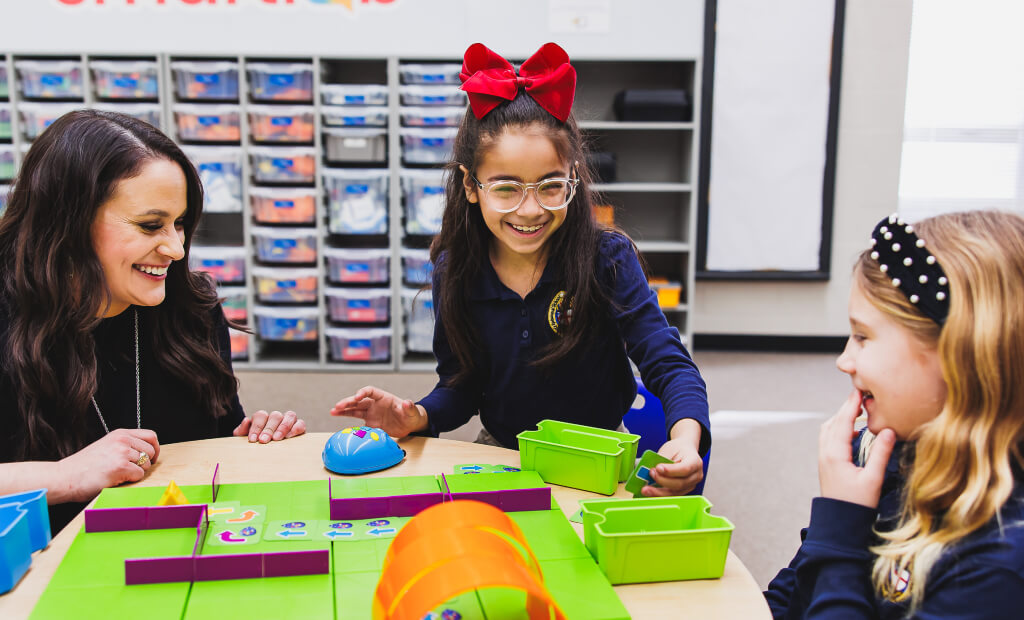 In a classroom, a woman and two girls enjoy playing with a toy, surrounded by educational materials.