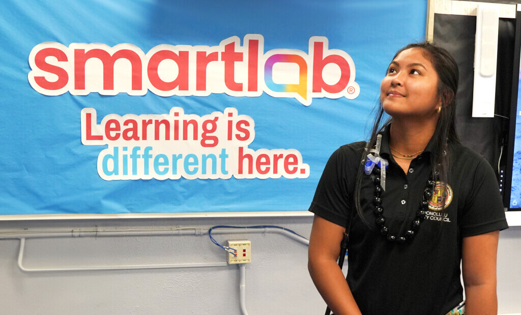 school girl in a black shirt with black hair looks up as she stands in front of a sign that reads smartlab learning is different here