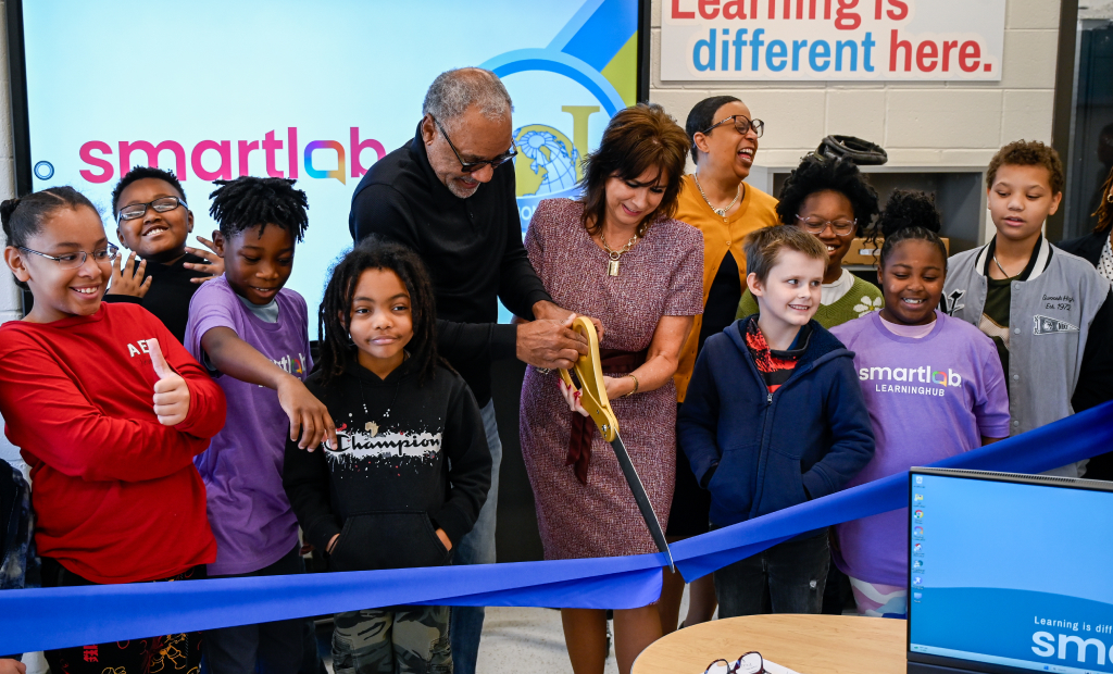 two adults cut a large blue ribbon with large scissors, school aged kids surround them