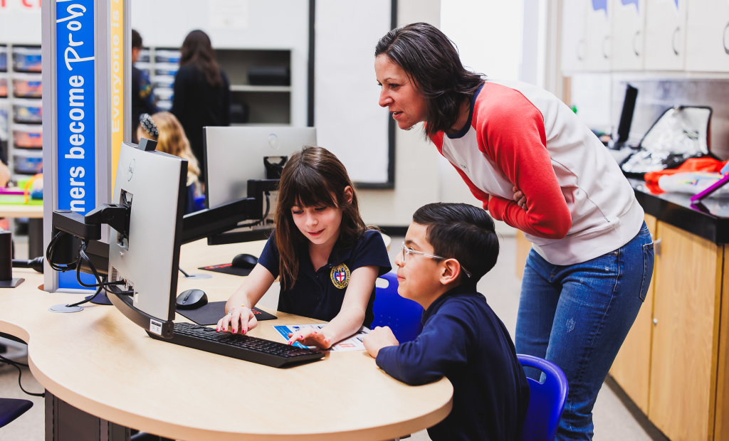a teacher leans over a computer station occupied by two school aged children in a smartlab setting