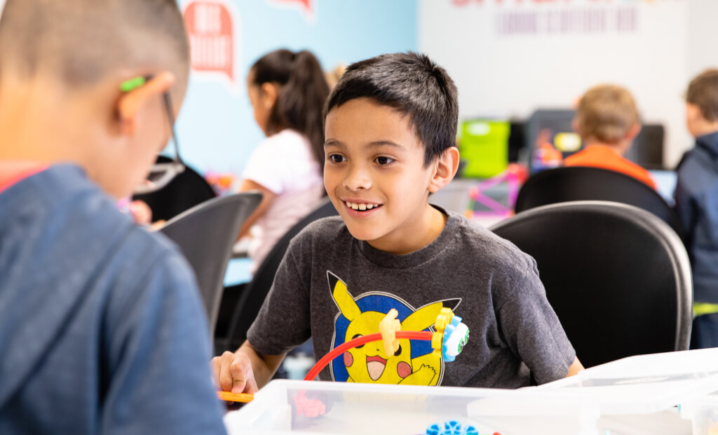 a school-aged boy with dark hair sits with fellow students at a table in a smartlab classroom setting