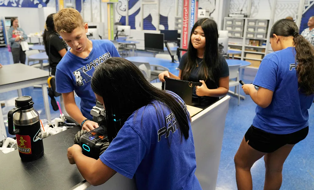Students in blue school shirts work with robotics equipment at a classroom table.