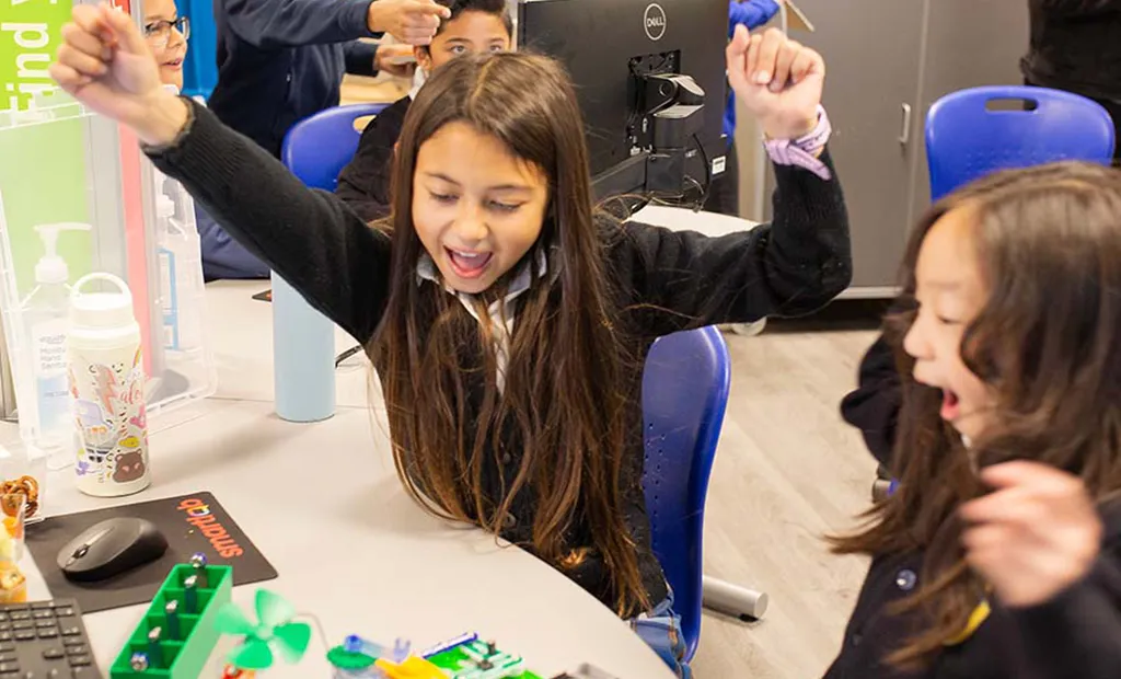 Girl with arms raised celebrating at a table with craft supplies and another child nearby.