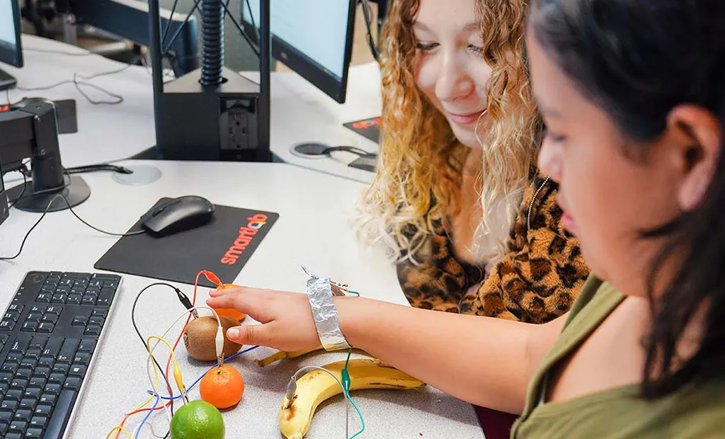 Two people examine a fruit-based electrical circuit project at a computer workstation.