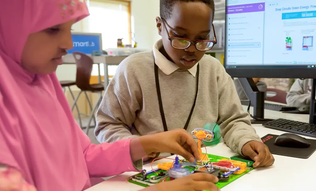 Two students examine a colorful circuit board learning kit at a desk in a classroom.